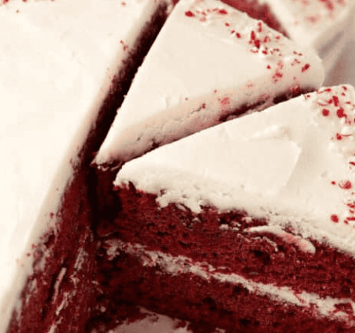 A red velvet cake with white frosting is sliced, revealing its rich red layers and creamy frosting. Perfect for those who love easy to make cakes, some slices are on a plate, and red sprinkles decorate the top edges.