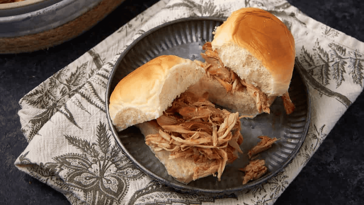 Two pulled pork sandwiches on soft white rolls are served on a round metal plate, placed on a patterned cloth napkin with a dark background—perfect for enjoying classic game day foods.