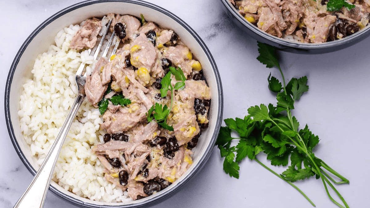 A bowl of white rice topped with a flavorful mix of tuna, black beans, corn, and cilantro—an easy twist inspired by crockpot recipes. A fork rests in the bowl and fresh cilantro is placed beside it on a light surface. Another bowl is partially visible.