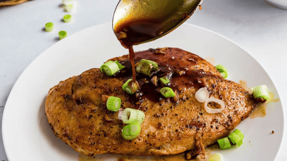 A spoon pours brown sauce over a seasoned, cooked chicken breast on a white plate, garnished with chopped green onions—one of the flavorful chicken breast recipes perfect for any meal.