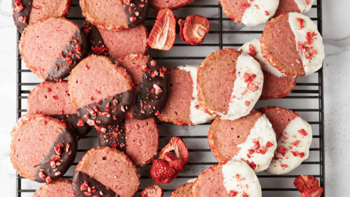 Round pink cookies, inspired by shortbread recipes, are dipped in dark or white chocolate and topped with crushed dried strawberries, arranged on a cooling rack with scattered strawberry pieces.