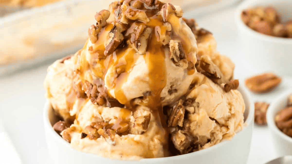 A bowl of creamy ice cream, made with sweetened condensed milk, is topped with chopped pecans and drizzled with caramel sauce, with extra pecans visible in the background—perfect for fans of desserts with sweetened condensed milk.