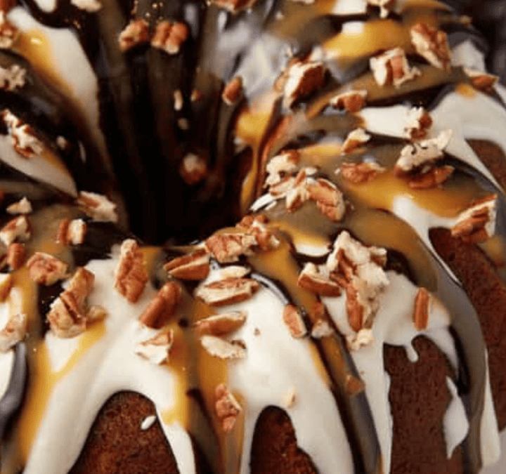 A bundt cake topped with white and caramel glaze, drizzled with chocolate, and sprinkled with chopped pecans, sits on a white plate—the perfect treat for a bit of baking therapy.