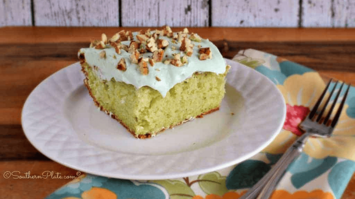 A slice of green cake, baked from a classic 9x13 cake pan, is topped with pale green frosting and chopped pecans on a white plate next to a fork and floral napkin. The background features a rustic wooden surface.