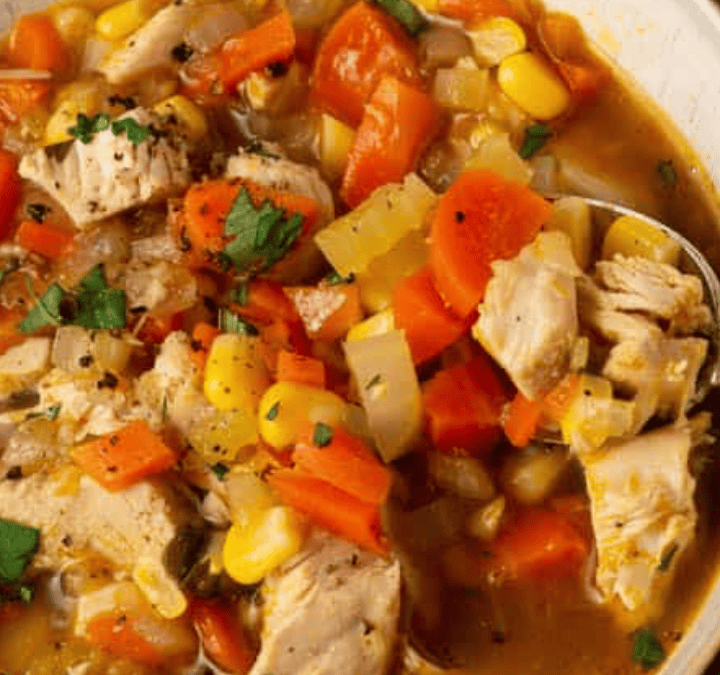 A close-up of a bowl of chicken stew, featuring chunks of chicken, carrots, corn, and celery in a brothy soup, garnished with chopped parsley and served with a spoon in the bowl.