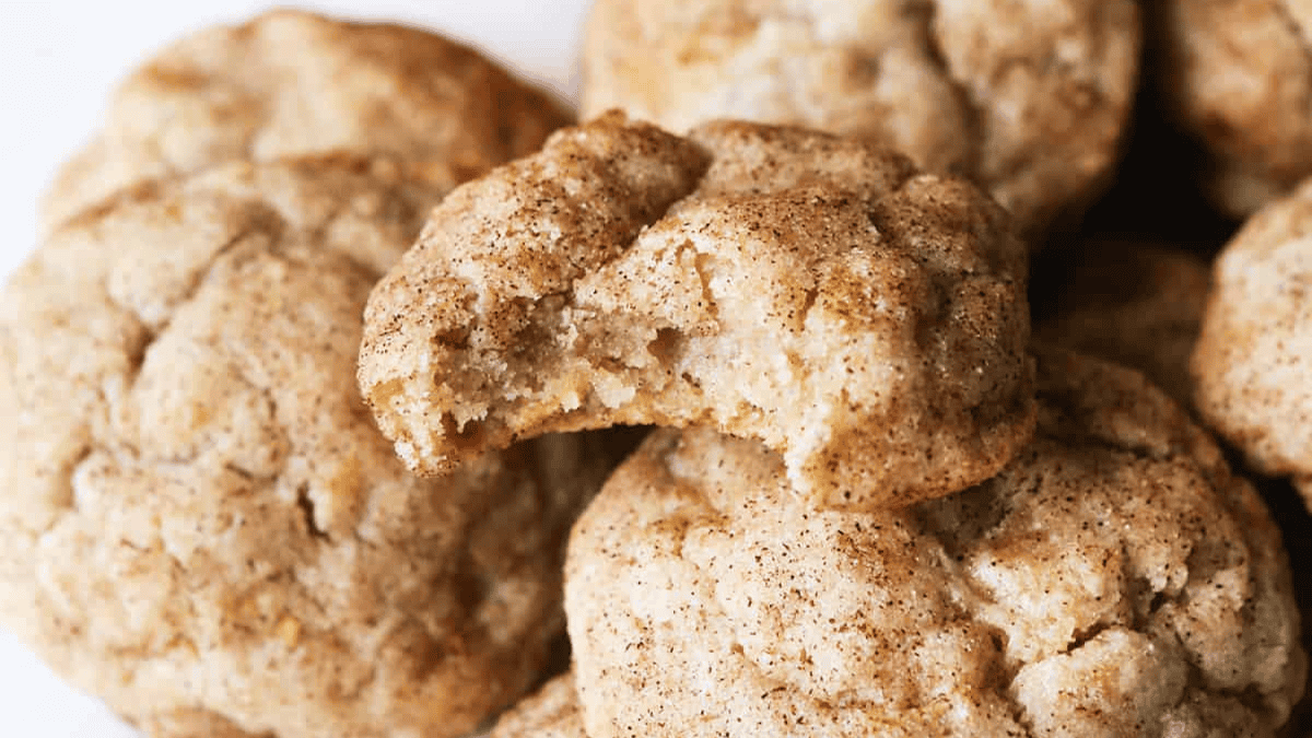 A close-up of several snickerdoodle cookies, made with cream of tartar, with one cookie broken in half to show its soft, fluffy interior. The cookies are coated in a cinnamon-sugar mixture and stacked closely together.