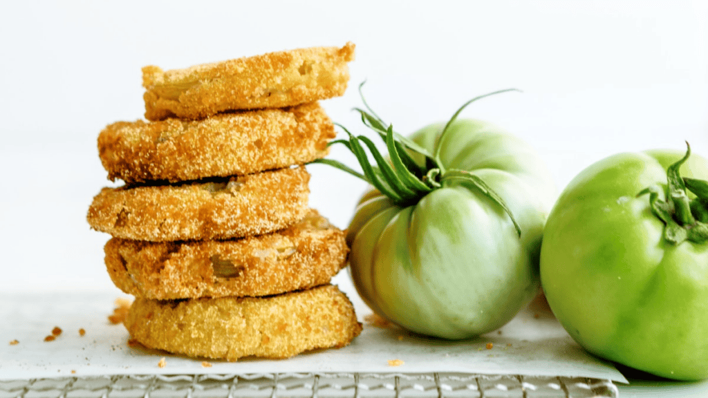 A stack of golden-brown fried green tomato slices, a classic choice for southern snacks, sits next to two whole green tomatoes on a light surface against a simple white background.