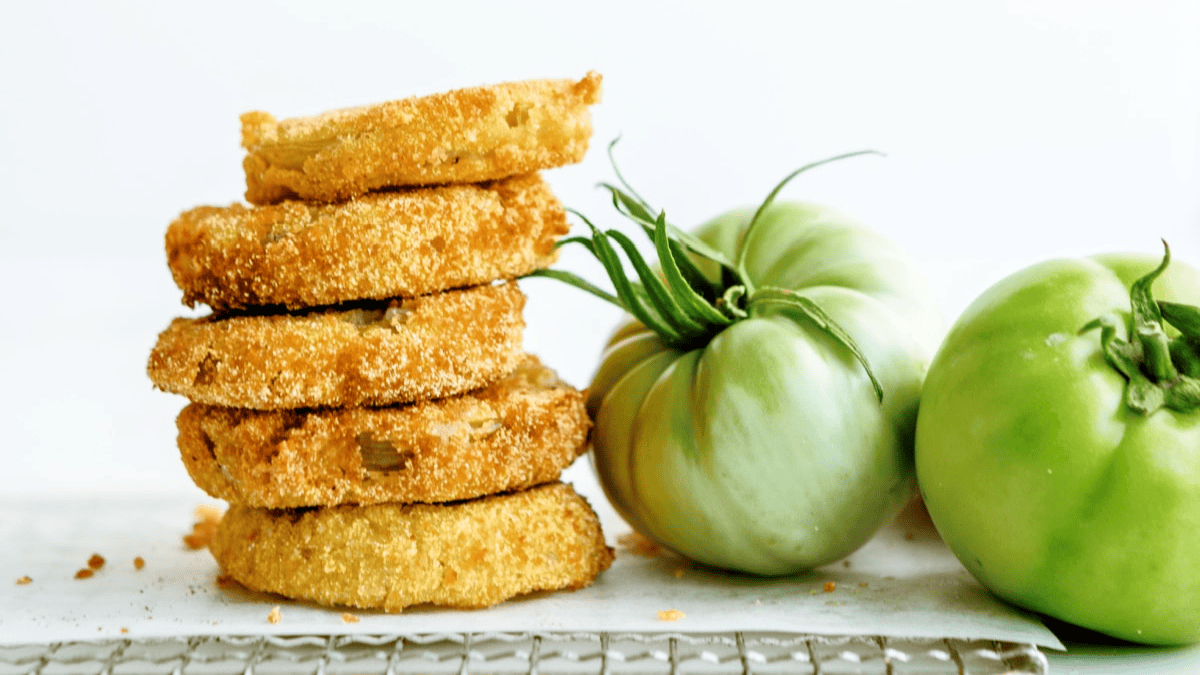 A stack of golden-brown fried green tomato slices, a classic choice for southern snacks, sits next to two whole green tomatoes on a light surface against a simple white background.