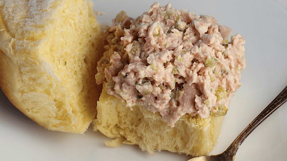 A soft, split dinner roll topped with creamy ham salad—complete with visible chopped pickles—is served on a white plate next to a small spoon, capturing the charm of old school recipes.