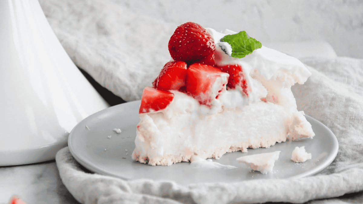 A slice of pavlova, made with a touch of cream of tartar, is topped with whipped cream, fresh strawberries, and a mint leaf on a gray plate, with a few crumbs scattered nearby.