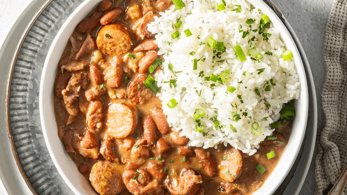 A bowl of red beans and sausage stew, one of the classic comfort foods, served with white rice and garnished with chopped green onions and herbs, placed on a plate with a beige napkin beside it.