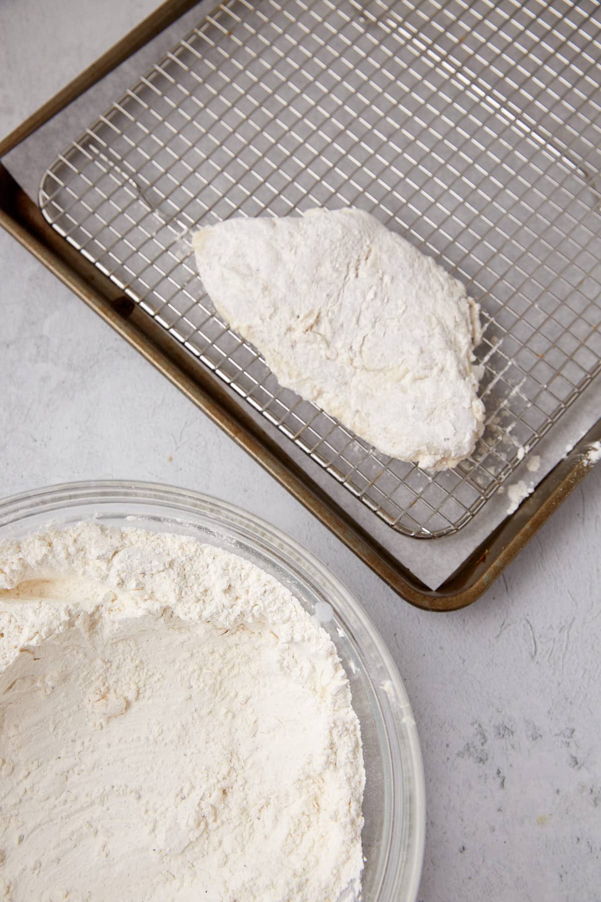 glass bowl filled with flour mixture sitting beside a baking sheet lined with a wire rack with a piece of the coated chicken on top