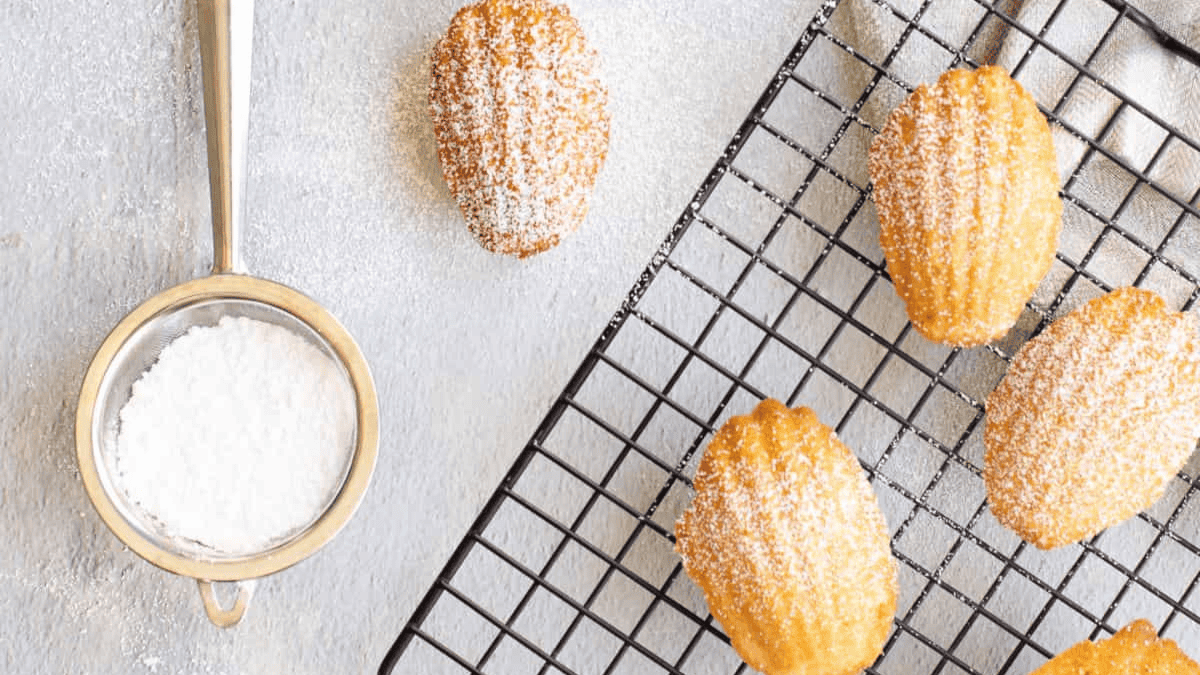 Golden madeleine cookies, a classic among French desserts, are dusted with powdered sugar and arranged on a cooling rack, with a metal sifter filled with powdered sugar placed nearby on a light-colored surface.