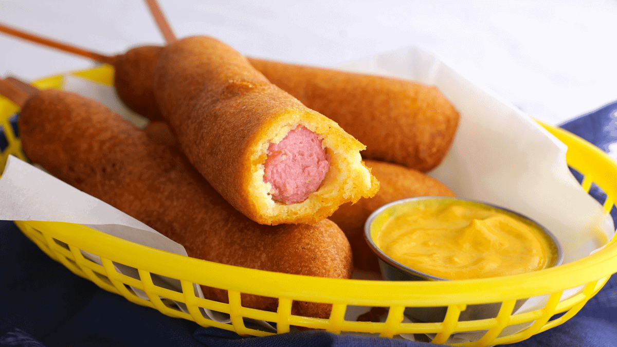 A yellow basket lined with white paper holds several corn dogs on sticks, perfect for air fryer recipes. One corn dog has a bite taken out, showing the pink sausage inside. A small metal cup of yellow mustard is beside them.