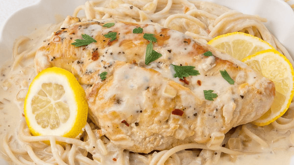 A creamy chicken breast served on a bed of pasta, garnished with fresh parsley and two lemon slices. Perfect for fans of pasta recipes, the rich sauce is seasoned with herbs and spices for a flavorful finish.