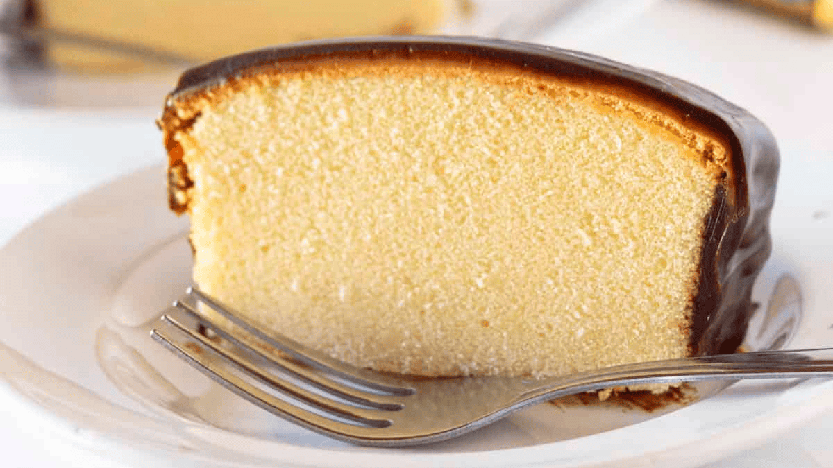 A slice of yellow pound cake with smooth chocolate icing on top, placed on a white plate with a silver fork resting in front—an inviting treat for lovers of classic pound cakes.