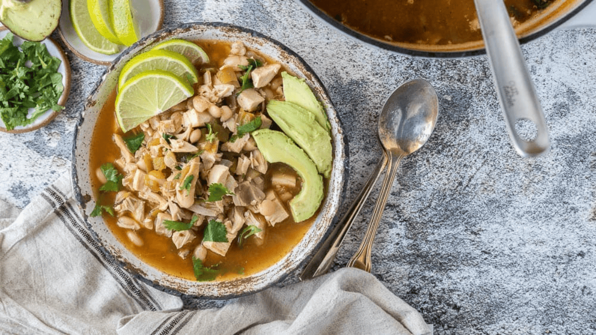 A bowl of soup with chunks of chicken, white beans, avocado slices, lime wedges, and cilantro—perfect for game day chili recipes. A spoon and napkin rest nearby, with fresh cilantro and lime on small plates beside the bowl.