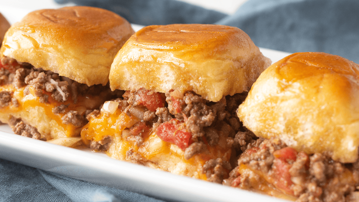 Three baked slider sandwiches filled with seasoned ground beef, diced tomatoes, and melted cheese are arranged on a white rectangular plate, showcasing one of the tastiest ground beef recipes, with a blue napkin in the background.