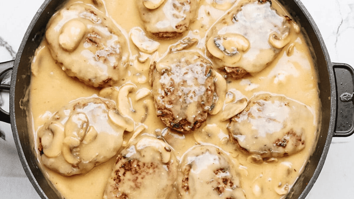 A pan filled with Salisbury steak patties covered in creamy brown mushroom gravy, with sliced mushrooms visible throughout the sauce—a true classic inspired by old school recipes.