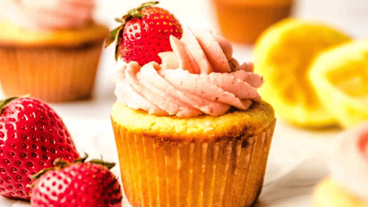 A close-up of valentine cupcakes topped with pink frosting and a fresh strawberry, with more cupcakes, strawberries, and lemon slices softly blurred in the background.
