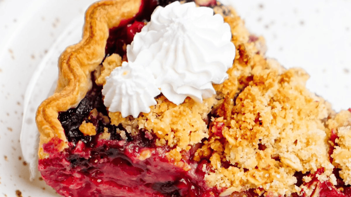 A close-up of a slice of berry crumble pie, featuring a flaky crust, crumbly topping, and whipped cream on top, served on a white plate—a perfect example of fruit infused recipes.