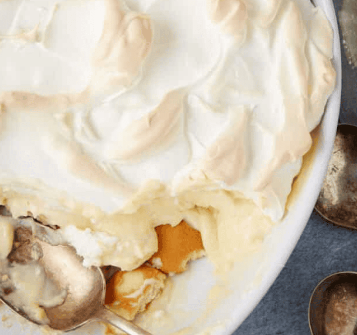 A classic southern desserts favorite, creamy banana pudding topped with fluffy meringue is partially served with a spoon, resting beside several vintage metal spoons on a gray surface.