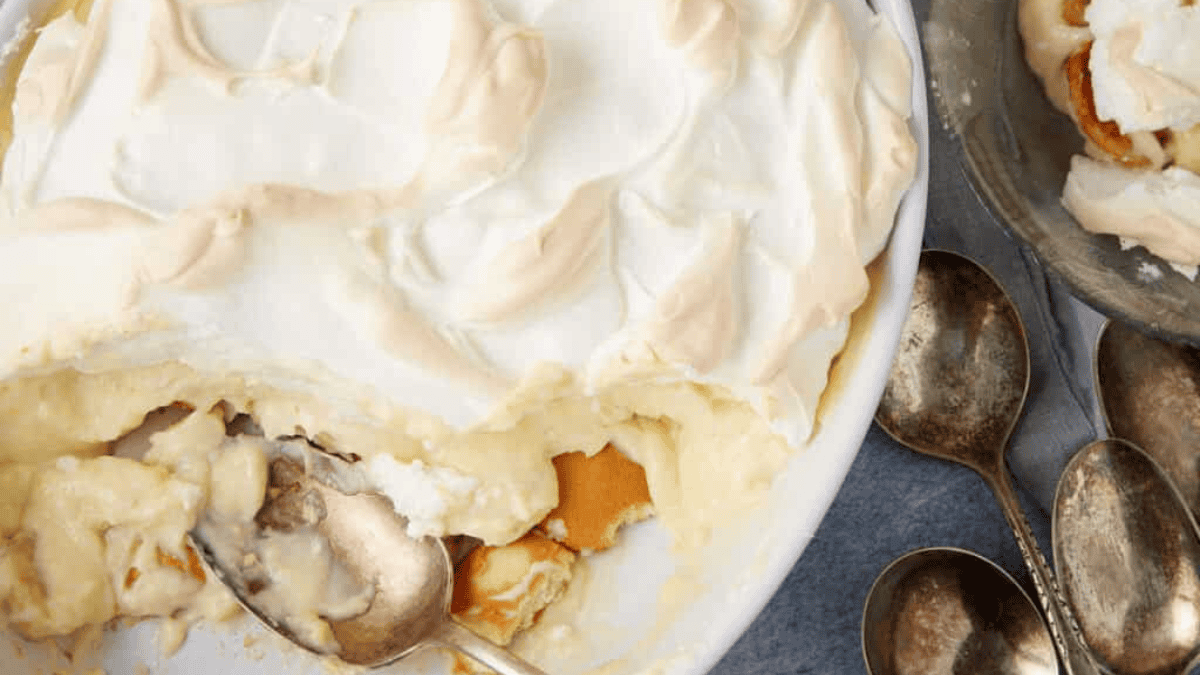 A classic southern desserts favorite, creamy banana pudding topped with fluffy meringue is partially served with a spoon, resting beside several vintage metal spoons on a gray surface.