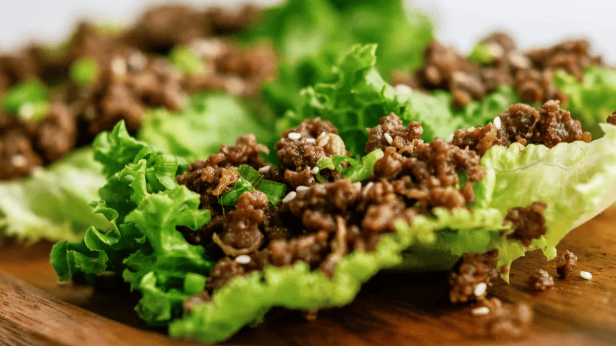 Savor one of the best ground beef recipes: seasoned ground beef served on fresh green lettuce leaves, garnished with sesame seeds atop a rustic wooden surface.