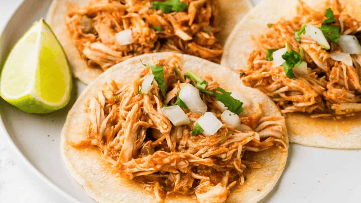 Three corn tortillas topped with shredded chicken, diced onions, and cilantro are served on a white plate with a lime wedge—perfect for fans of easy slow cooker recipes.