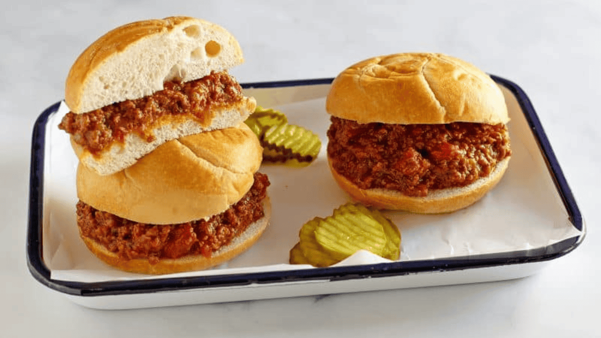 Two sloppy joe sandwiches on round buns sit on a white tray with a blue rim, accompanied by pickles. One sandwich is stacked atop the other on parchment paper—classic comfort food perfect for slow cooker recipes.