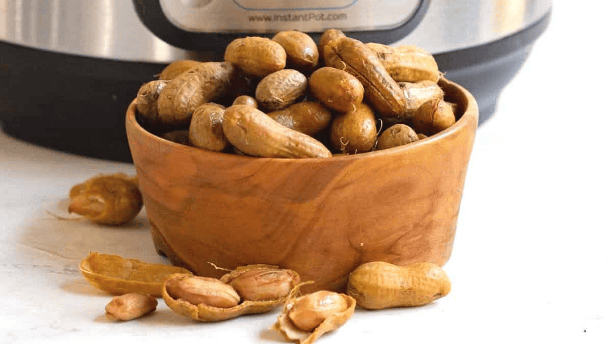 A wooden bowl filled with boiled peanuts, a beloved southern snack, sits on a white surface with scattered peanut shells and loose peanuts nearby. A kitchen appliance is blurred in the background.