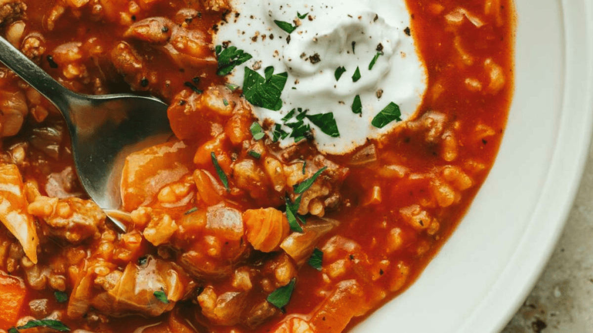 A close-up of a bowl of tomato-based stew with ground meat, rice, and vegetables—one of those comforting soups—topped with a dollop of sour cream and chopped herbs. A spoon is resting inside the bowl.