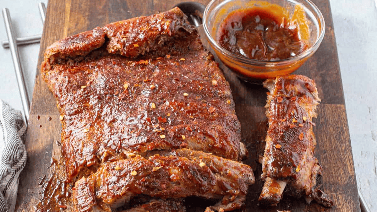 A slab of cooked ribs with a crispy, seasoned crust sits on a wooden board beside a glass bowl of barbecue sauce, with a few ribs cut and separated—one of the most mouthwatering pork recipes for any barbecue lover.