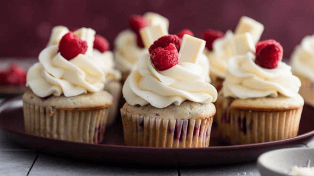A plate of Valentine cupcakes topped with swirls of white frosting, fresh raspberries, and pieces of white chocolate, arranged on a dark plate with a blurred background.