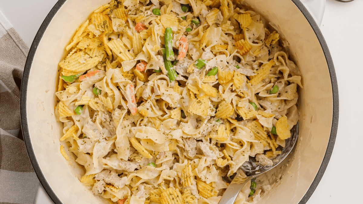 A large white pot filled with creamy noodle casserole, perfect for weeknight dinners, topped with crumbled potato chips, sliced green onions, and mixed vegetables, with a serving spoon on the side.