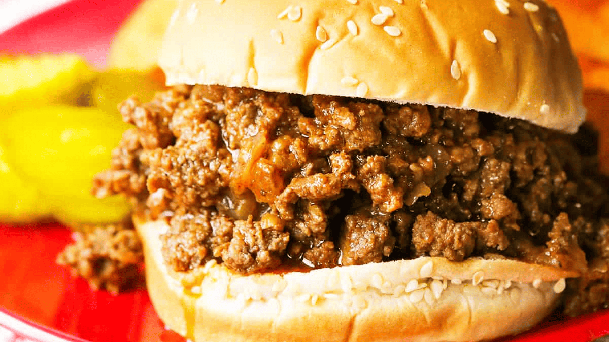 A close-up of a sloppy joe sandwich with seasoned ground beef on a sesame seed bun, served on a red plate with pickle slices—classic old school meals at their best.