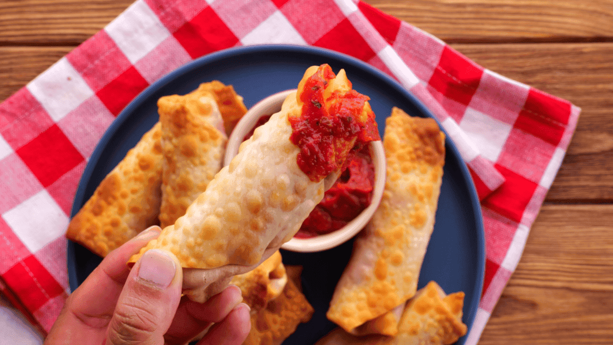 A hand holds a crispy, golden egg roll from an air fryer recipe, dipped in red marinara sauce, with more egg rolls on a blue plate and a red-and-white checkered napkin underneath.