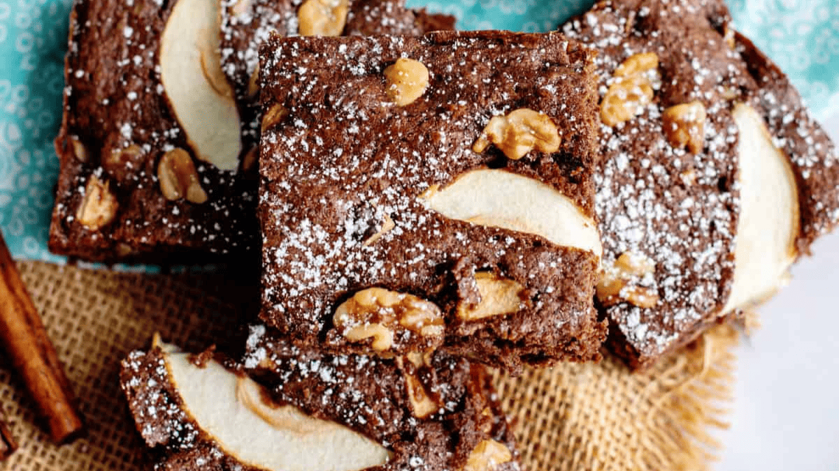 Chocolate brownies topped with powdered sugar, sliced apples, and walnuts—an irresistible fusion of apple desserts—arranged on burlap fabric with a cinnamon stick nearby.