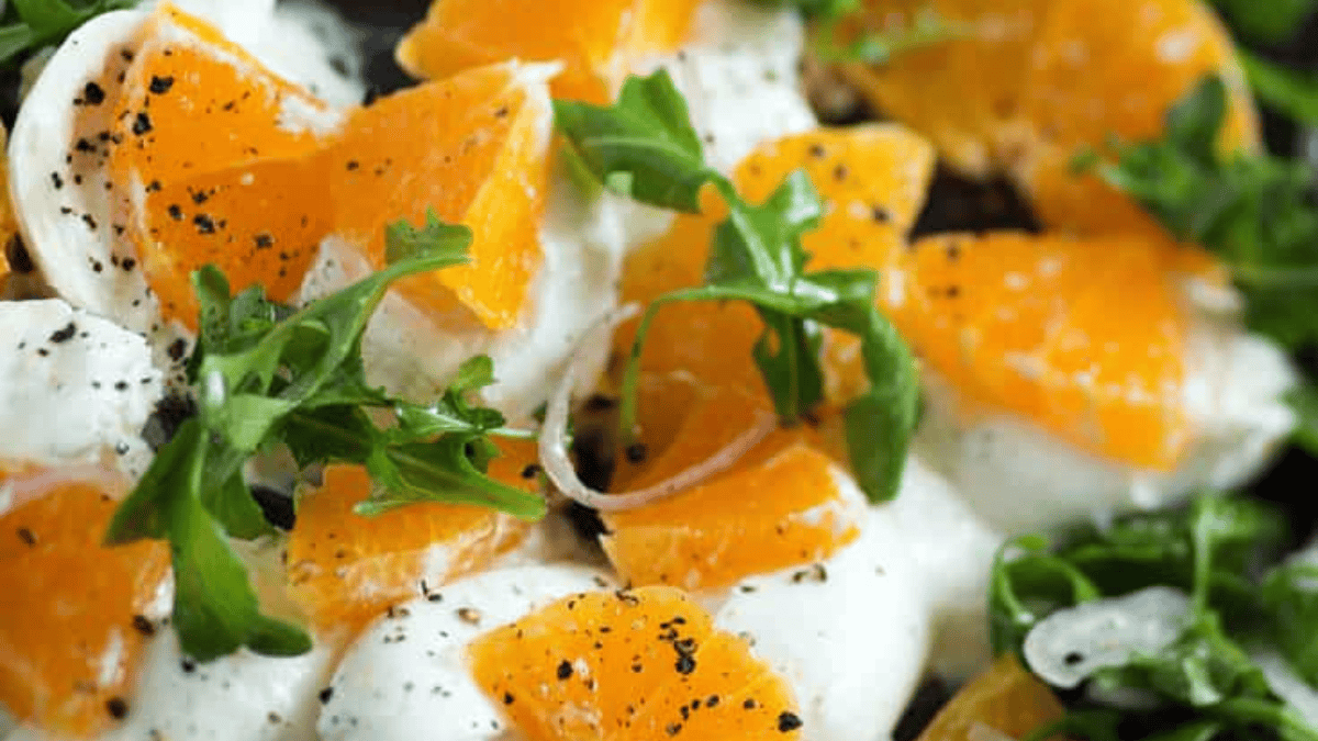 Close-up of a vibrant spring salad with slices of fresh mozzarella, orange segments, arugula leaves, and cracked black pepper sprinkled on top.