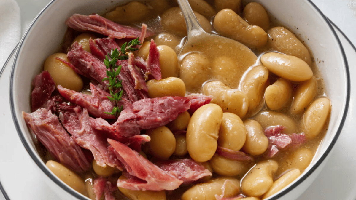 A bowl of cooked lima beans in a broth, topped with shredded ham and garnished with a sprig of fresh thyme, evokes the comforting flavors of old school meals, with a spoon resting in the bowl.