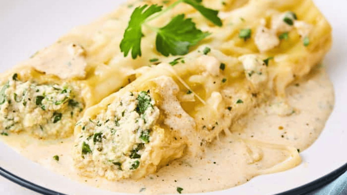 Two veggie packed stuffed cannelloni pasta rolls topped with creamy white sauce and garnished with fresh herbs, served on a white plate.