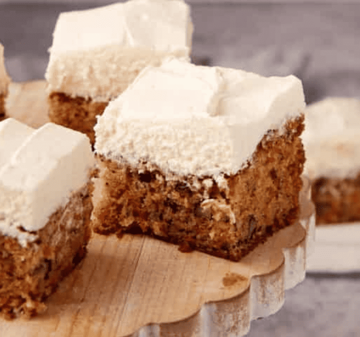 Squares of carrot cake with thick, creamy white frosting are arranged on a wooden cake stand, making this one of the best spring desserts. Additional pieces sit invitingly on white plates in the background.