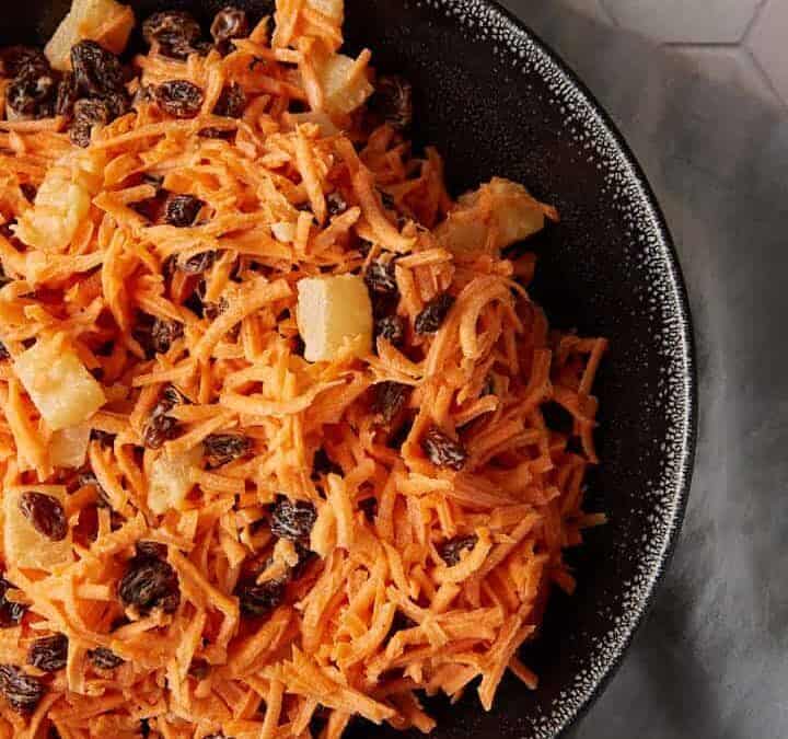 A bowl of carrot and raisin salad next to a bunch of carrots.