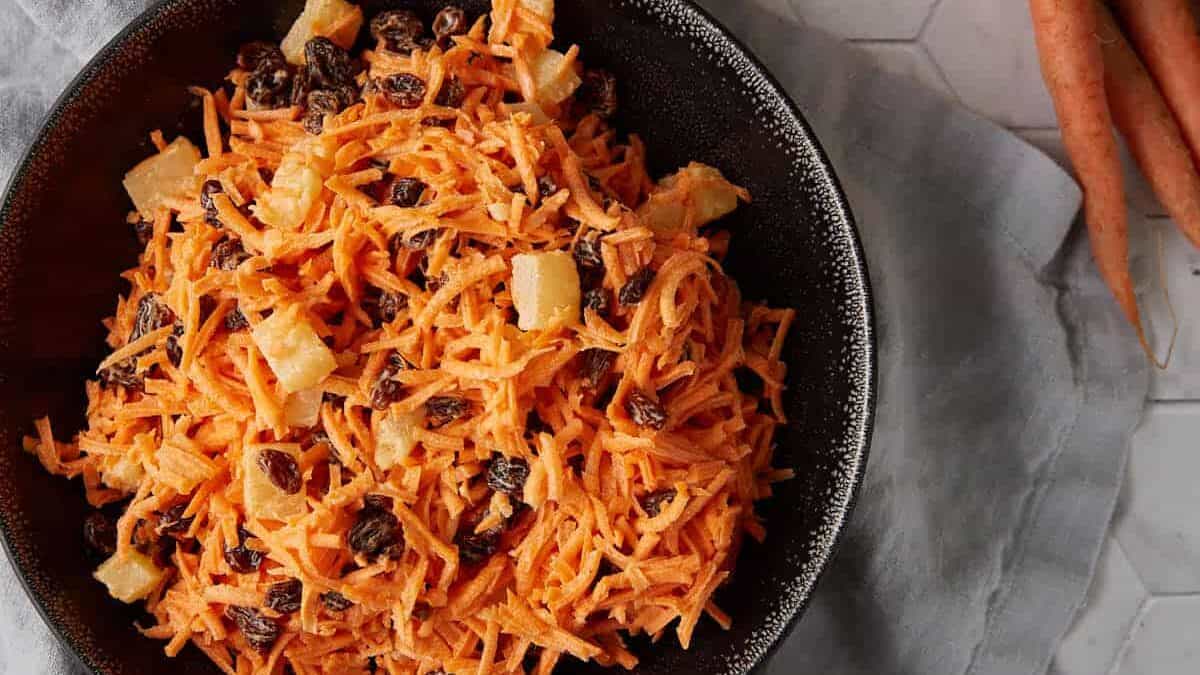 A bowl of carrot and raisin salad next to a bunch of carrots.
