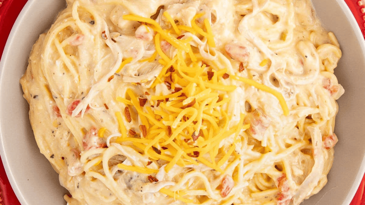 A bowl of creamy spaghetti topped with shredded cheddar cheese and small pieces of tomato—perfect for cozy slow cooker dinners.