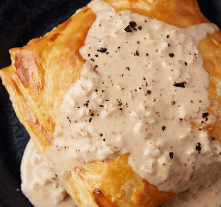 A golden-brown pastry square topped with creamy white sauce and cracked black pepper, served on a dark plate—perfect for easy meals.