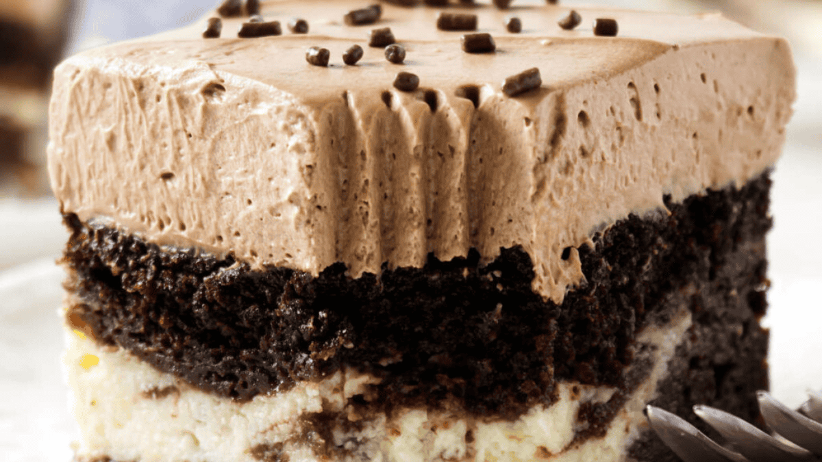 A close-up of a layered dessert shows creamy chocolate frosting, a thick layer of dark chocolate cake, and a white cheesecake base, topped with chocolate sprinkles. A fork is partially visible beside this indulgent example of layered desserts.