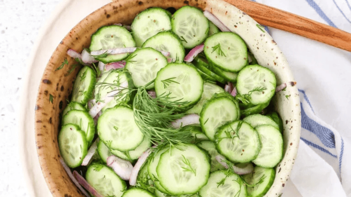 A bowl filled with sliced cucumbers, thin red onion slices, and fresh dill makes a perfect choice for spring salads. A wooden spoon rests beside the bowl, with part of a blue-striped cloth napkin visible.