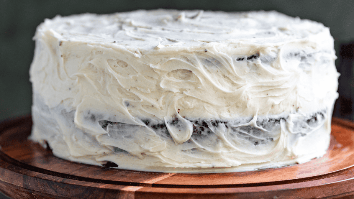A round layer cake with white, textured frosting is displayed on a wooden cake stand. Some dark chocolate cake layers are faintly visible through the frosting, making it a stunning example of effortless desserts.
