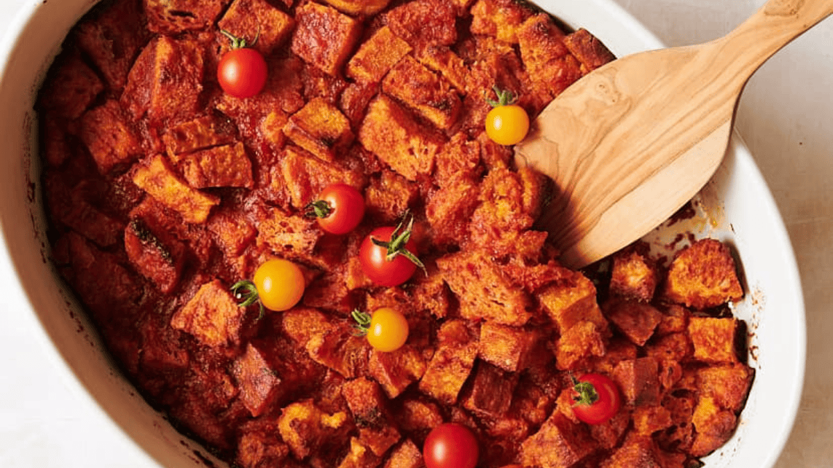 A baked dish of cubed bread in a tomato-based sauce, topped with red and yellow cherry tomatoes, is served in a vintage casserole dish with a wooden serving spoon. Perfect for those who love the charm of vintage dishes.